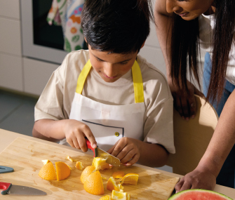 Un niño con delantal cortando trozos de naranja en una tabla de cortar de madera bajo la supervisión de un adulto.