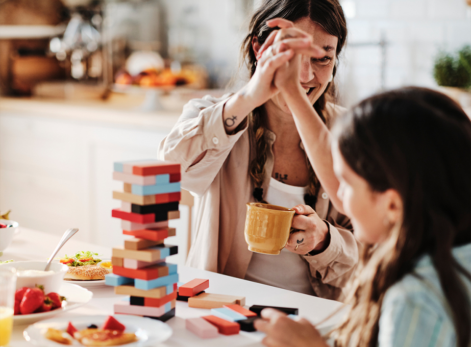 Una mujer y una niña chocan las manos celebrando mientras juegan con una torre de bloques de colores en una mesa de desayuno.