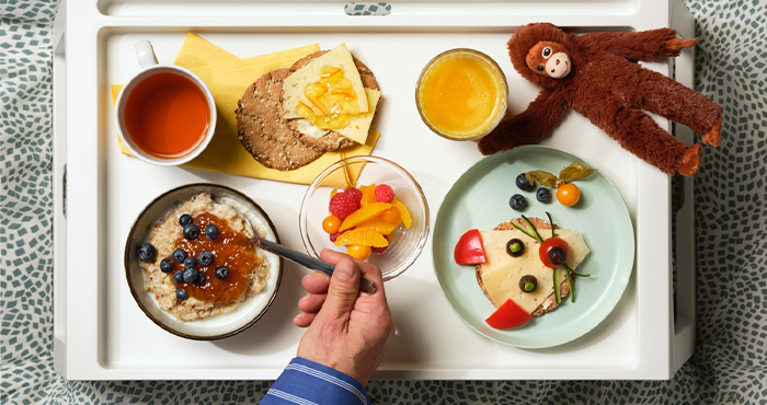 Vista cenital de una bandeja de desayuno con avena, fruta fresca, zumo y una tostada decorada con cara de animal junto a un peluche de mono.