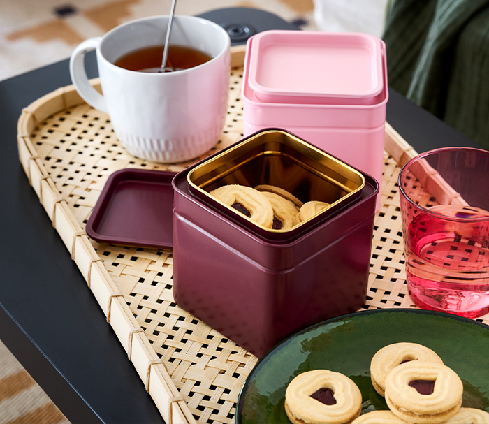 Una bandeja de mimbre con una taza de té, una lata rosa cerrada y una lata granate abierta llena de galletas con forma de corazón, con más galletas en un plato verde al lado.