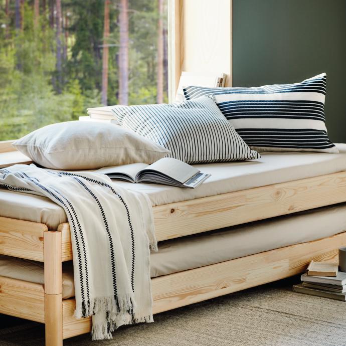 Cama nido apilable de madera de pino con cojines de rayas, una manta blanca y un libro abierto, situada junto a un gran ventanal con vistas al bosque.