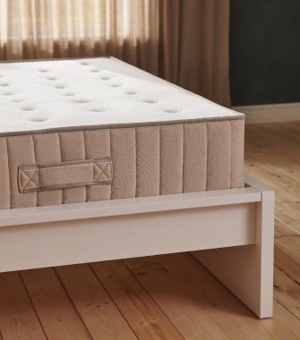 Primer plano de un colchón grueso de color beige con asa lateral, apoyado sobre una estructura de cama de madera blanca minimalista.