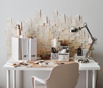 Escritorio blanco de oficina en casa con una lámpara plateada, organizadores y un gran collage artístico de recortes de papel en la pared trasera.