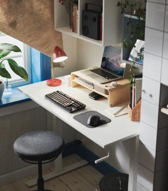 Espacio de trabajo ergonómico en casa que cuenta con un escritorio elevable blanco, un portátil sobre un soporte de madera, un teclado mecánico y una lámpara de escritorio roja.