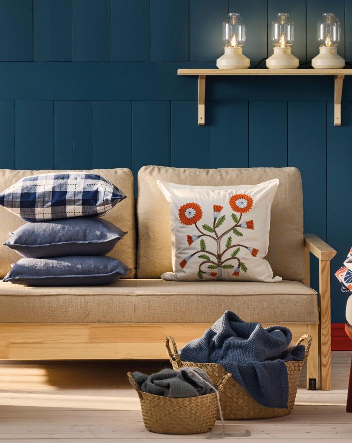 Living room with beige sofa, stacked blue cushions, decorative floral pillow, woven baskets, and wall shelf with lamps.