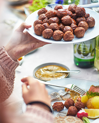 A hand holding HUVUDROLL meatballs on a plate, and another plate with prepared SILL DILL, alongside a DRAGON cutlery set.