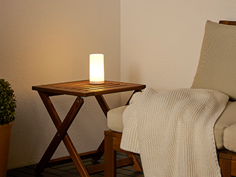 SOLVINDEN decorative lamp on a NÄMMARÖ folding table, beside a NÄMMARÖ modular sofa with a HUMLEMOTT blanket on top.