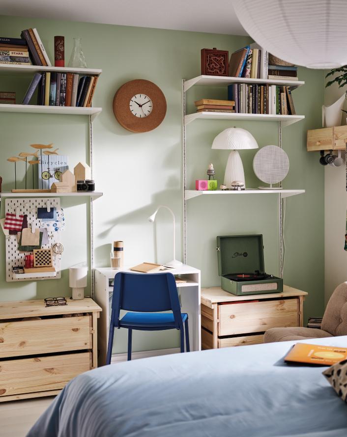 Cozy room with light green walls, a small desk, white shelves filled with books and decor, and a bed in the foreground.