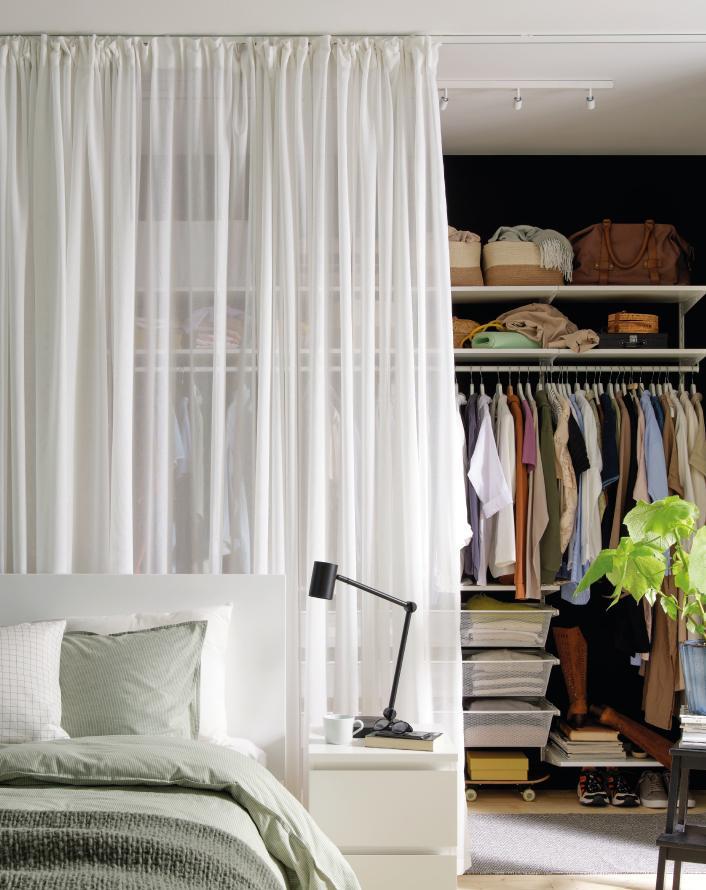 Modern bedroom with sheer white curtains separating the bed from an open closet featuring hanging clothes, storage boxes, and minimalist decor.