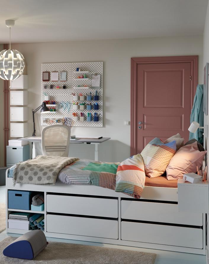 Youth bedroom with a white bed featuring built-in storage, a workspace desk, a wall organizer board, and colorful bedding.