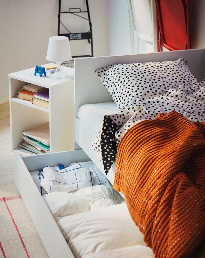 Modern bedroom with a white bed, black polka dot bedding, an orange knitted blanket, and an open storage drawer underneath the bed.