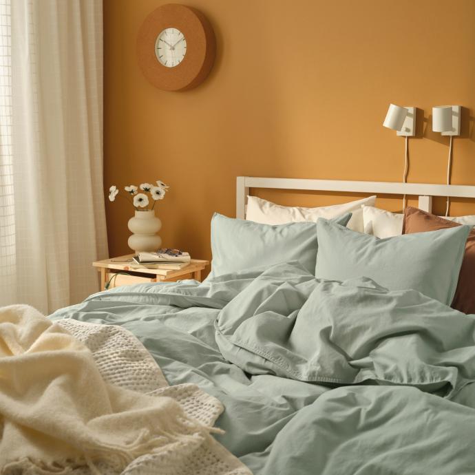 Un dormitorio acogedor con ropa de cama verde salvia, una manta crema y una pared amarillo mostaza con un reloj de madera.