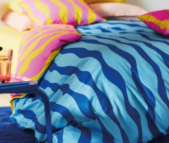 Primer plano de una cama con ropa de cama vibrante que incluye patrones ondulados en azul y cojines en rosa y amarillo.