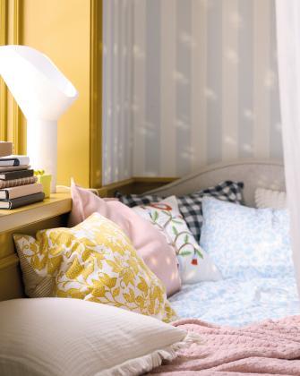 Un rincón de cama acogedor con múltiples cojines decorativos en tonos rosa, azul y amarillo junto a una pared de rayas.