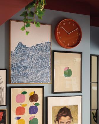 Una pared de galería con cuadros de olas azules y manzanas de colores sobre una pared azul junto a un reloj naranja.