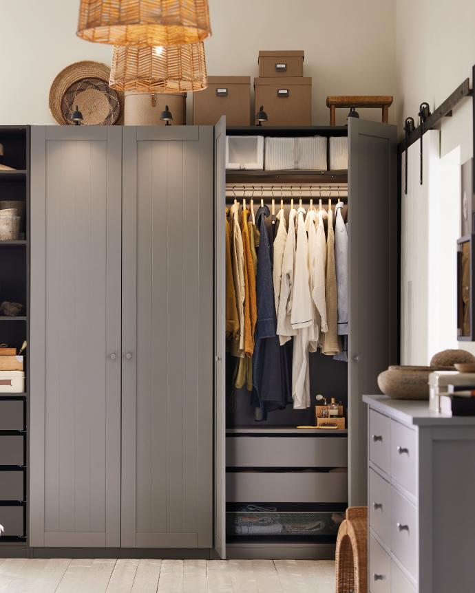 A stylish gray wardrobe with open doors revealing hanging clothes, storage boxes, and drawers in a cozy, organized bedroom.