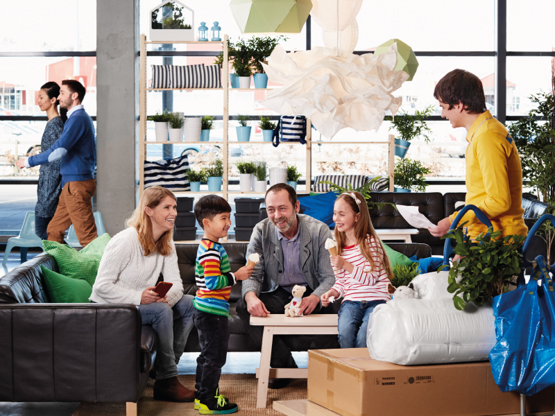A family sits on a showroom sofa interacting with an IKEA employee in a yellow shirt. The children hold ice cream, while boxes and a blue shopping bag sit in the foreground.