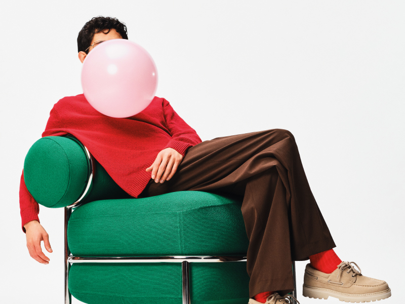 A person in a red sweater lounges on a green modern armchair, blowing a large pink bubble gum bubble against a white background.