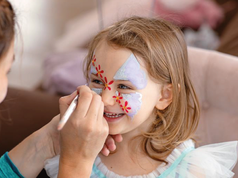 Close-up of a smiling young girl getting her face painted with a colorful butterfly and flower design.