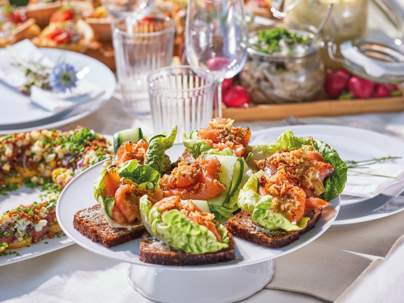 A white tiered stand filled with smoked salmon open-faced sandwiches on dark rye bread, topped with lettuce, cucumber, and crispy onions on a festive table.