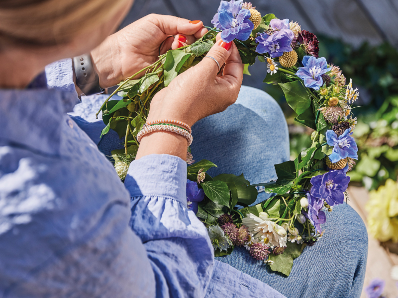 Close-up of a person's hands weaving blue, white, and purple flowers into a green leafy crown resting on their lap.
