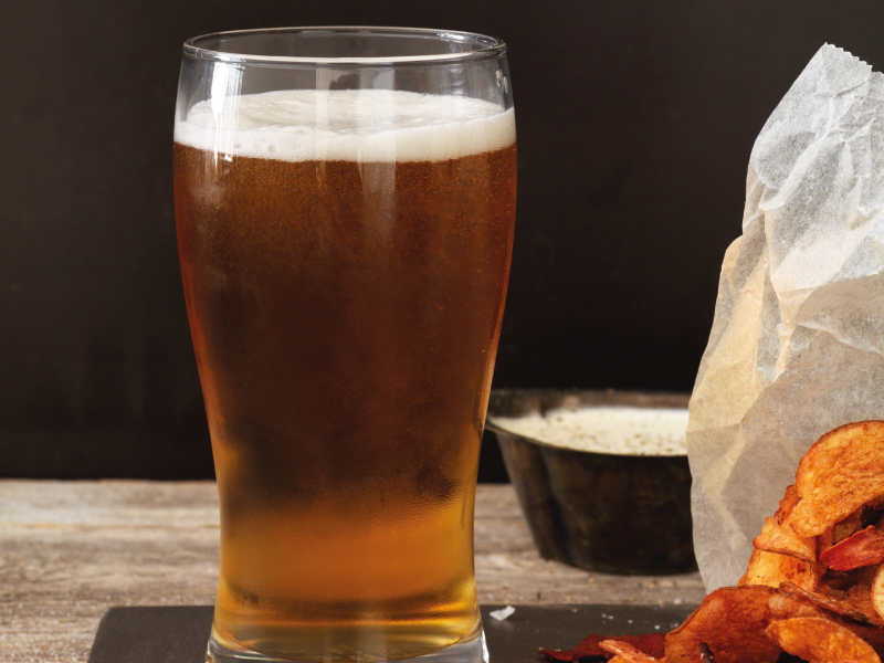 A pint glass filled with amber beer and white foam stands next to a bag of potato chips and a small bowl of dip on a rustic wooden surface.