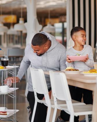 Una persona adulta sirviendo comida mientras un niño sonriente sostiene un tazón en un comedor iluminado.