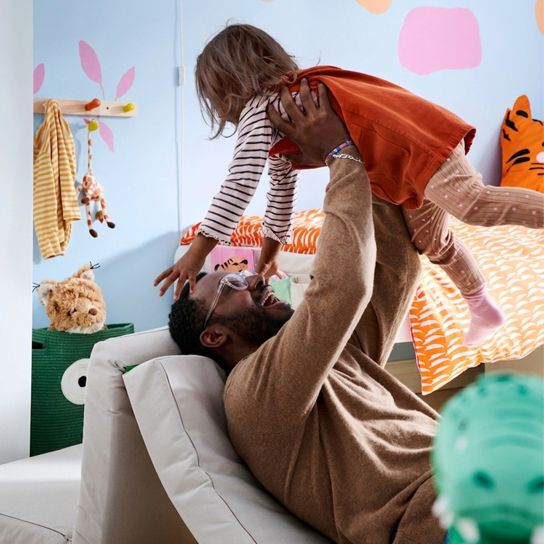 A laughing father lies back on a modular foam play couch while playfully lifting his young child up in the air inside a colorful children's bedroom.