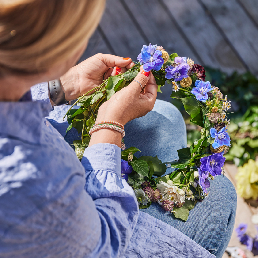 An over-the-shoulder view of a woman weaving a traditional midsummer floral crown with green leaves, blue, and white flowers on her lap.