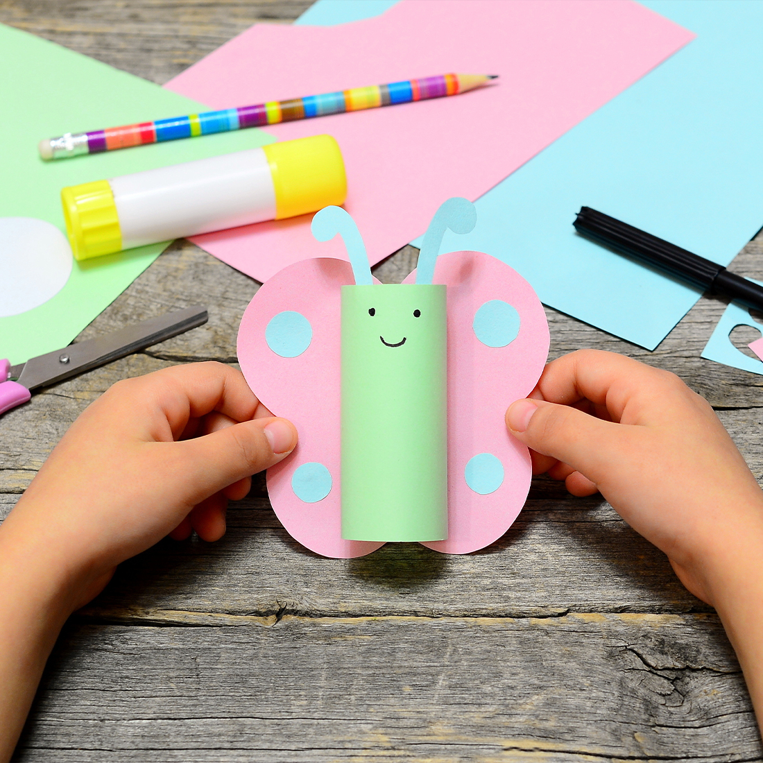 A child's hands holding a handmade butterfly craft made from a green paper roll and pink wings, surrounded by crafting supplies on a wooden table.