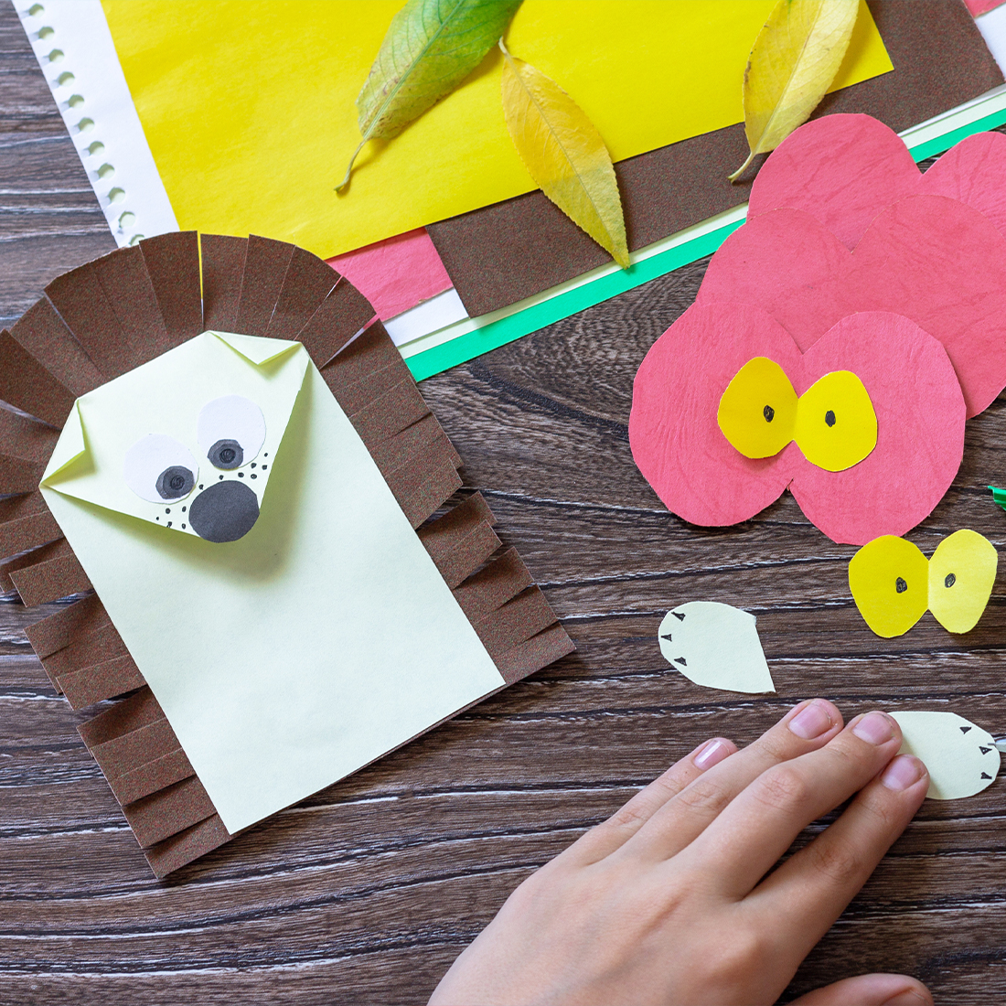 A top-down view of a person making paper animal crafts on a wooden table, featuring a finished paper hedgehog, red paper cutouts, and yellow eyes.