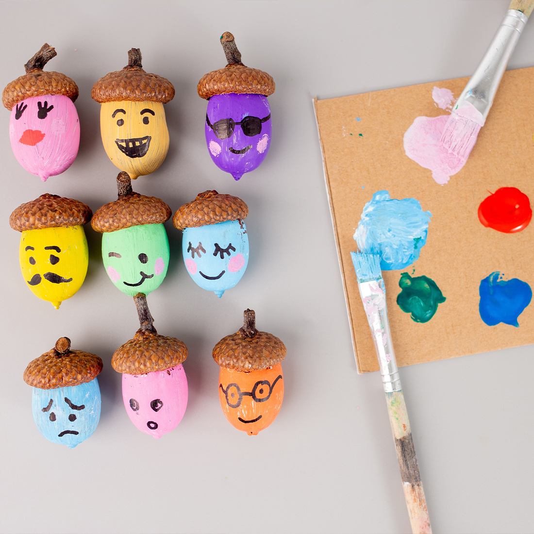 A top-down view of nine colorful acorns painted with various funny faces, arranged in rows next to a cardboard palette with paint and brushes.