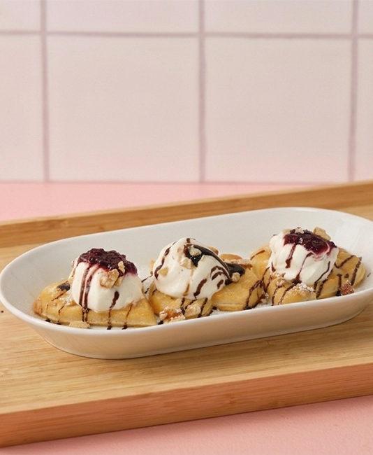 Three heart-shaped waffles topped with vanilla ice cream, berry jam, and chocolate syrup, served in an oval white dish on a wooden tray.