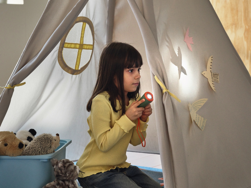 A young girl sitting inside a grey play tent, holding a small flashlight and shining it to create bird-shaped shadows on the fabric.