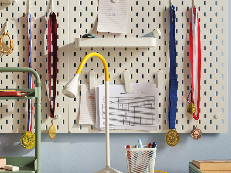 An organized white pegboard over a desk, holding school papers, notes, and several sports medals hanging from hooks, next to a yellow desk lamp.