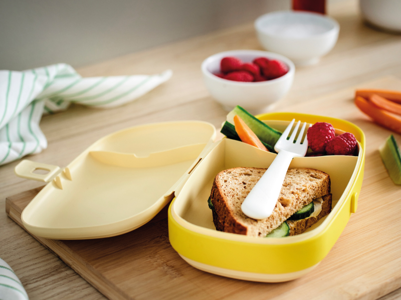 An open yellow lunchbox on a wooden cutting board, filled with a whole-grain sandwich, cucumber and carrot sticks, raspberries, and a white fork.