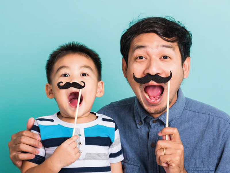 A man and a young boy smiling joyfully while holding fake prop mustaches over their mouths against a light blue background.