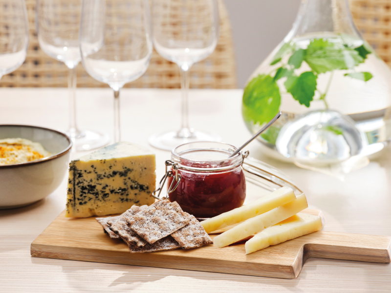 A wooden board with blue cheese, cheese sticks, crackers, and a jar of jam, with empty wine glasses in the background.