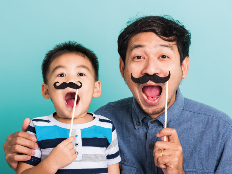 Father and son posing playfully holding fake mustaches over a light blue background.