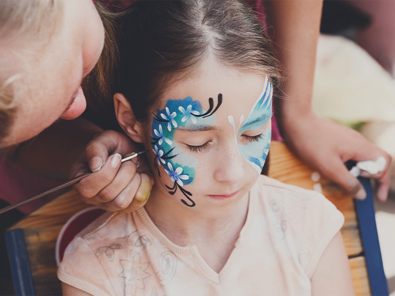 Artist painting a blue butterfly and flower design on a young girl's face.
