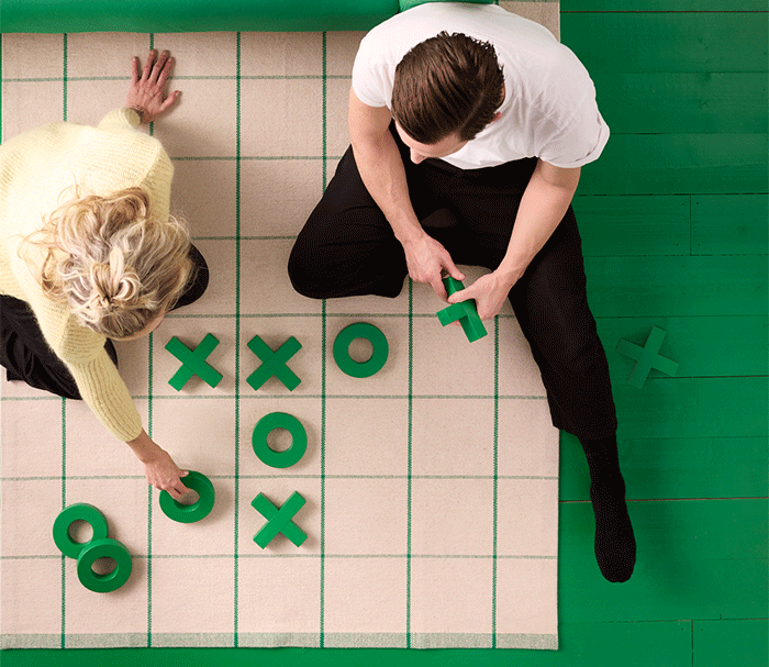 Overhead view of a couple playing giant tic-tac-toe with green pieces on a rug.