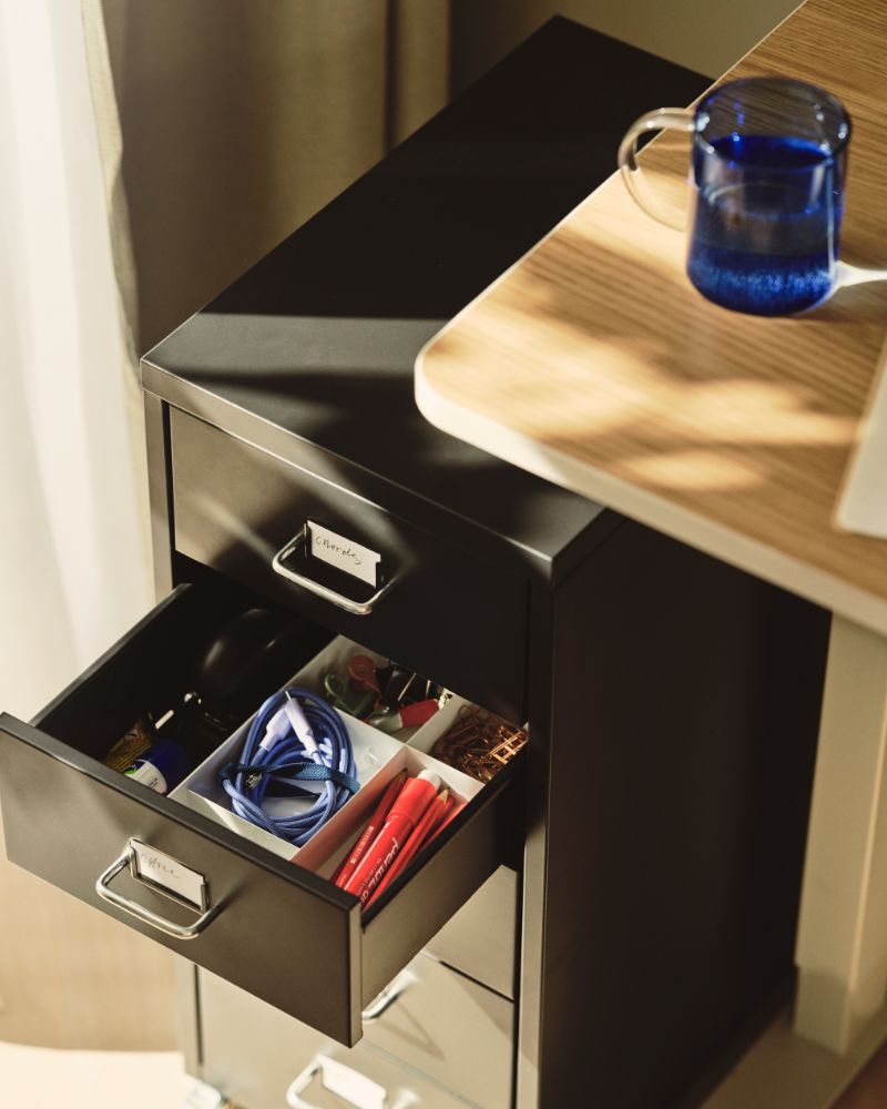 A close-up of a black HELMER drawer unit on castors with one drawer open, showing neatly arranged items inside.