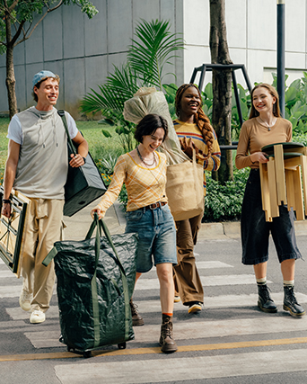 Group of people walking outside carrying bags, a potted plant, and wooden stools.