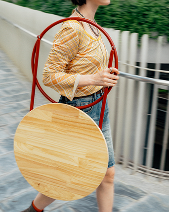 Person walking while carrying a round wooden folding table with red legs.