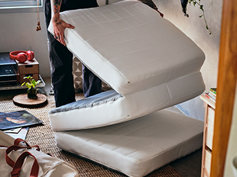 Person unfolding a white folding mattress on a rug inside a room.