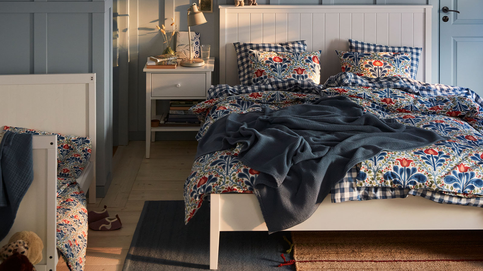 A blue bedroom with a white GULLABERG bed with floral bed linen, a blue throw and a white bedside table.