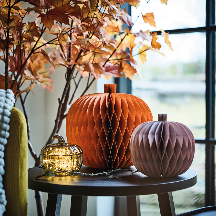 Calabazas decorativas de papel sobre una mesa junto a hojas otoñales y una ventana.