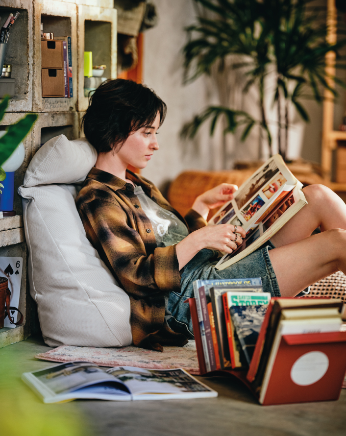 Person sitting on the floor reading a book in a corner with plants and shelving.