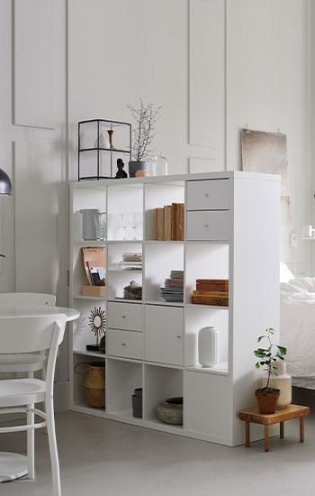 Un dormitorio en el que destaca una estantería KALLAX blanca llena de objetos decorativos y cajas de almacenamiento, junto a una cama de matrimonio con sábanas blancas.