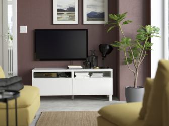 White TV console with a television, lamp, and books against a mauve wall, framed by a fiddle-leaf fig tree and yellow chairs.